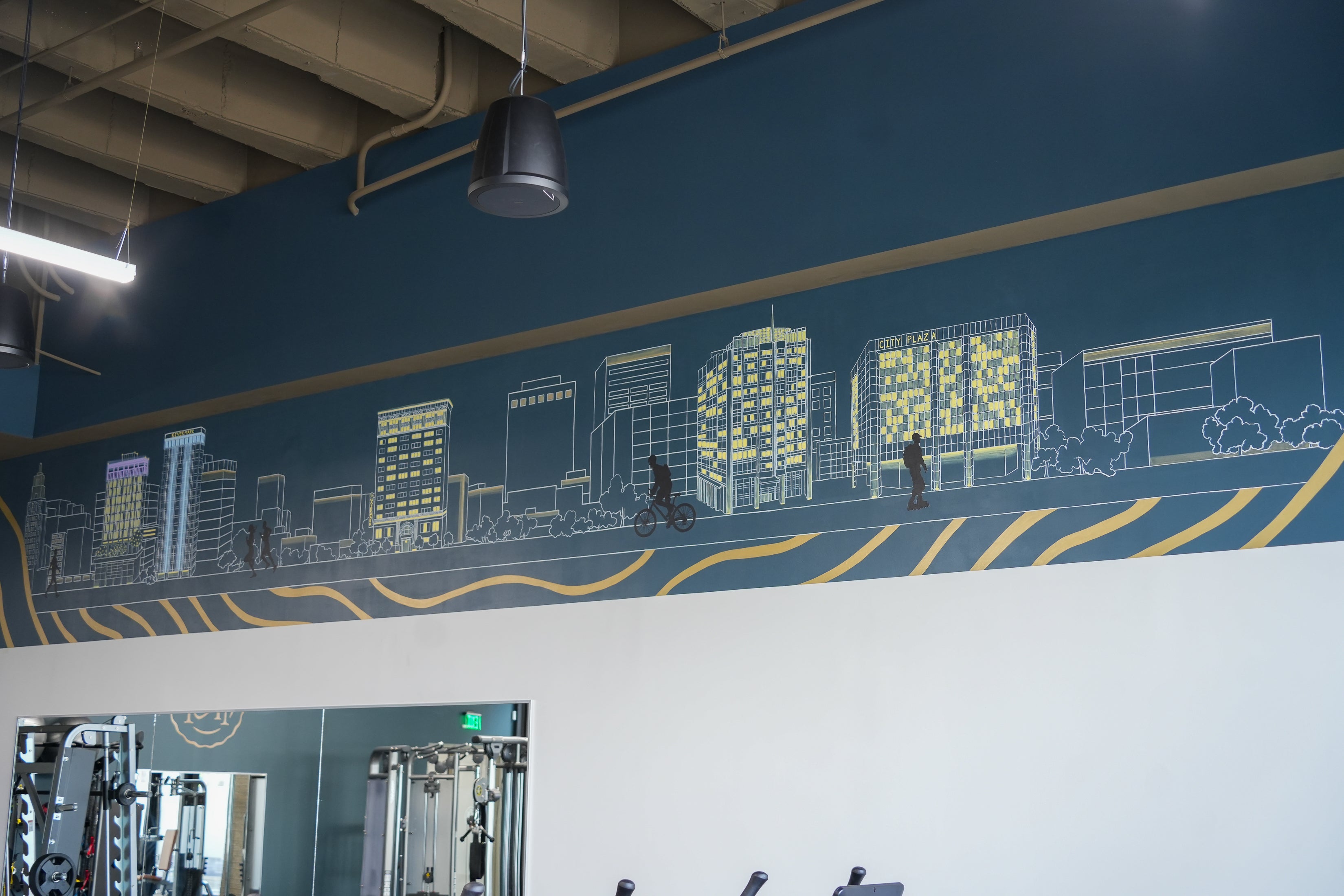 A large wall mural depicting a stylized city skyline with buildings, pedestrians, and cyclists, painted on a dark blue wall in a gym interior.