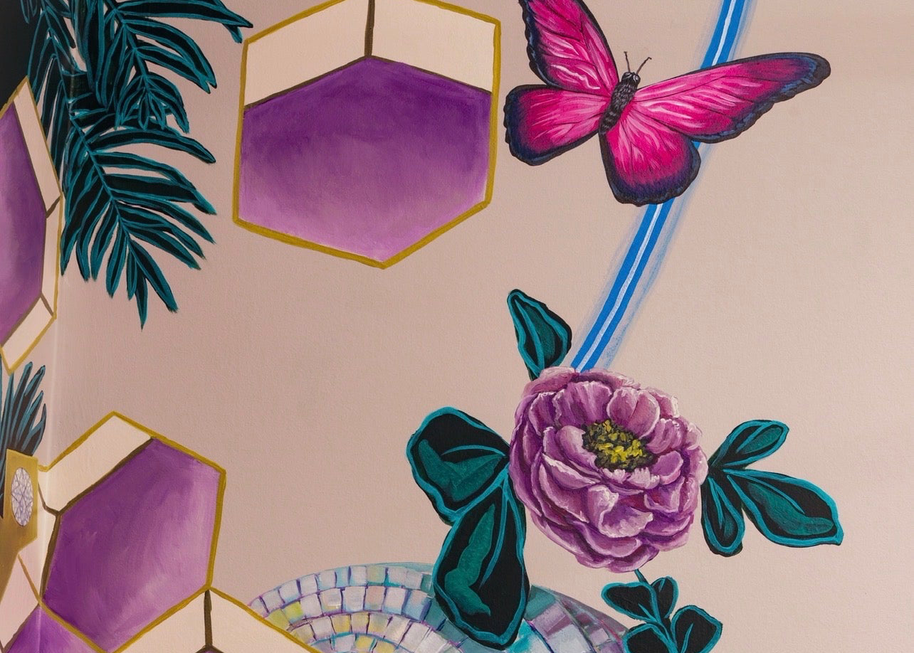 A close-up of a decorative surface featuring purple geometric hexagons outlined in gold, a pink butterfly, a purple flower, dark teal leaves, and a circular mosaic tile motif on a pale blush background.